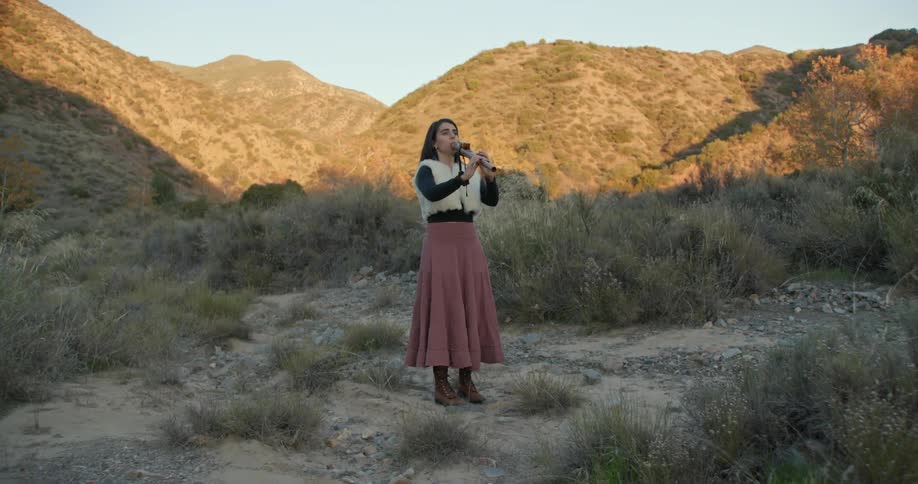 A woman wearing a long skirt is stanidng outside playing a clarinet and there are mountains in the distance