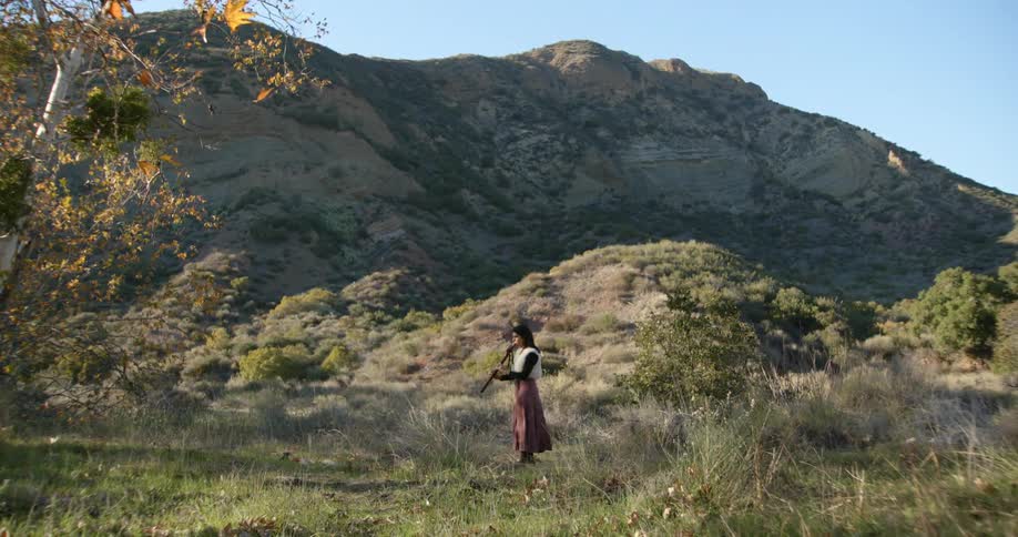 Woman wearing white vest and brown skirt plays bassoon in the field near a hill