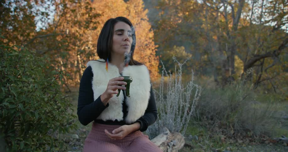 Woman sitting over a log outdoors holds a censer burning aromatic incense at evening