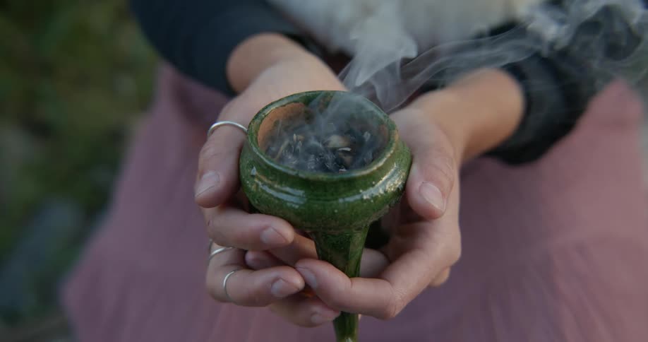 Close up of white smoke coming out of an incense burner on the hands of a woman sitting in the grass
