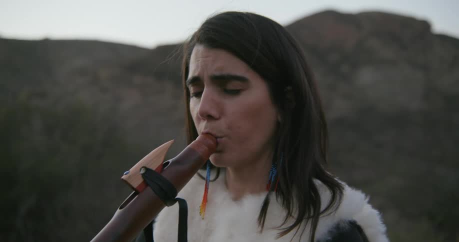 Woman wearing a long skirt plays a long wooden flute on a valley in the mountains at morning