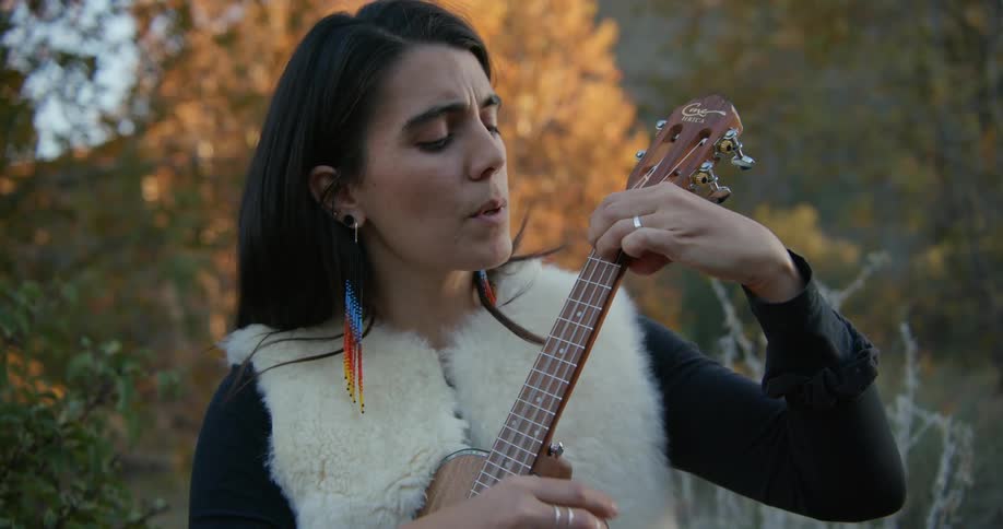 Woman with dark hair colorful earrings white vests and black shirt plays ukulele near the autumn forest singing a song