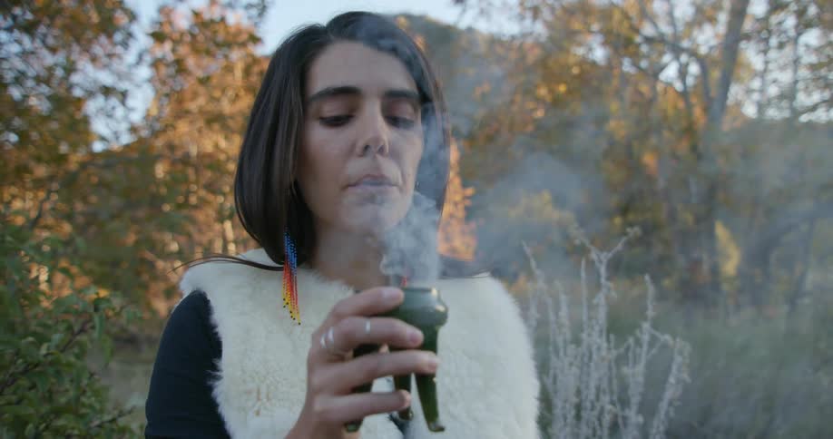 A woman is standing outside holding an incense burner and smelling the smoke and she holds her arm out and turns her head