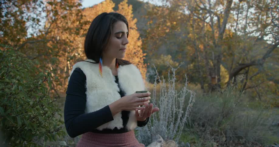 Woman sitting over a log outdoors holds a censer burning aromatic incense at evening
