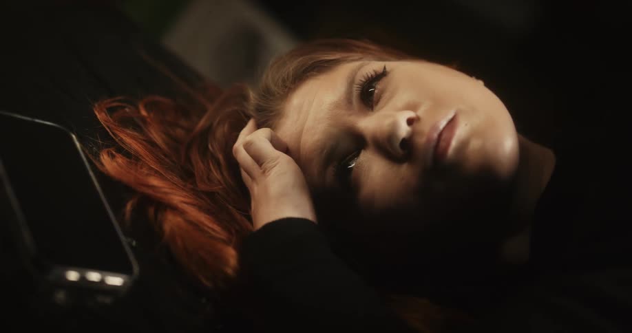 Light shines on the face of a red haired woman as she lies down with her phone next to her