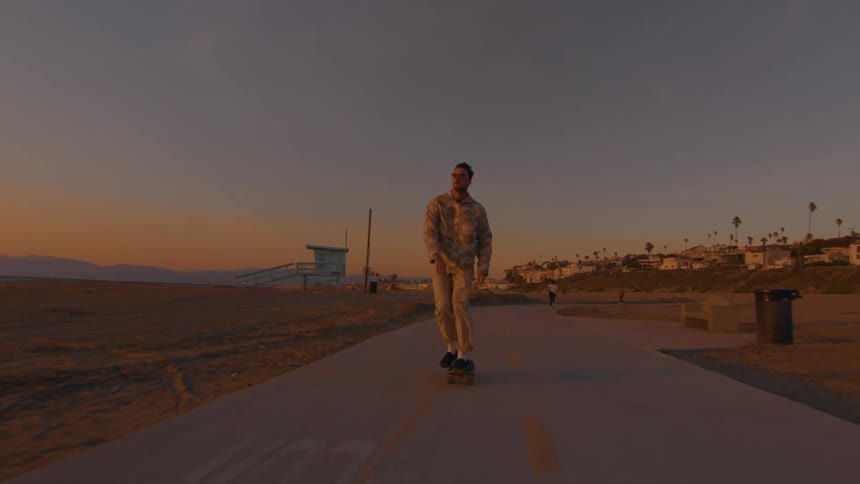 A man is riding a skateboard on a road along the beach and there are buildings in the distance