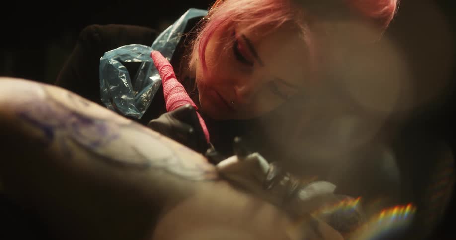 A female tattoo artist looks closely as she draws a tattoo on a person