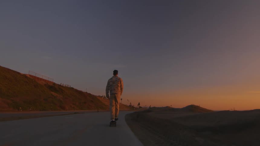 A man is filmed from behind riding a skateboard down a concrete road