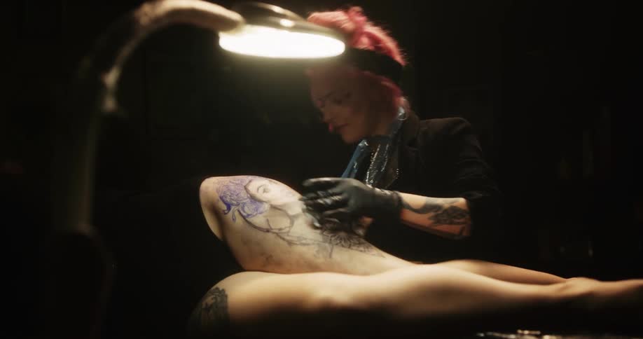 Happy tattoo artist with colorful hair tattooing on the leg of a customer