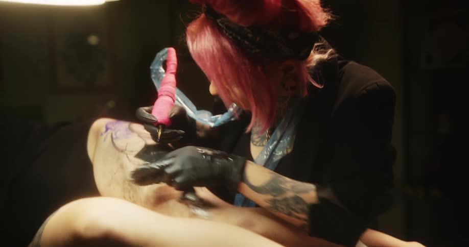 A woman tattoos another womans thigh who is laying down on a table
