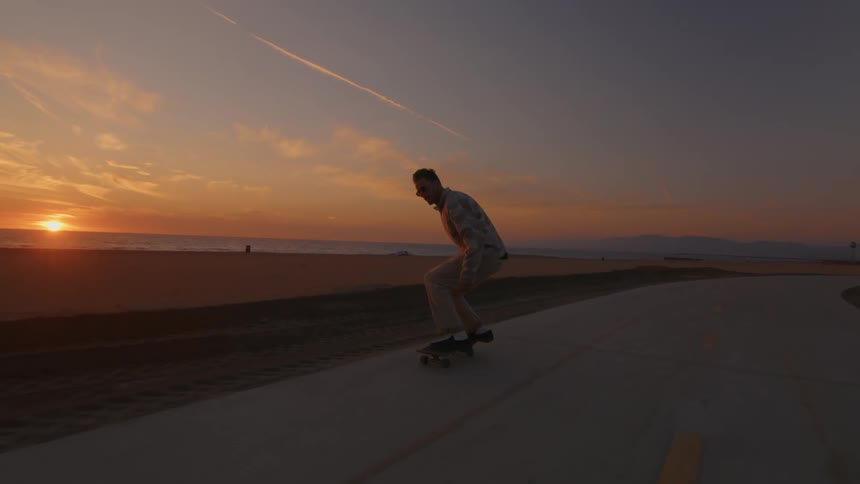 A man is riding a skateboard on a road along the coast and he leaps into the air with the sun setting in the distance