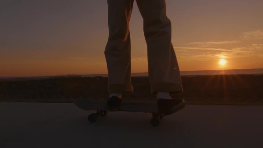 A man is riding a skateboard on a road along the coast and he leaps into the air with the sun setting in the distance