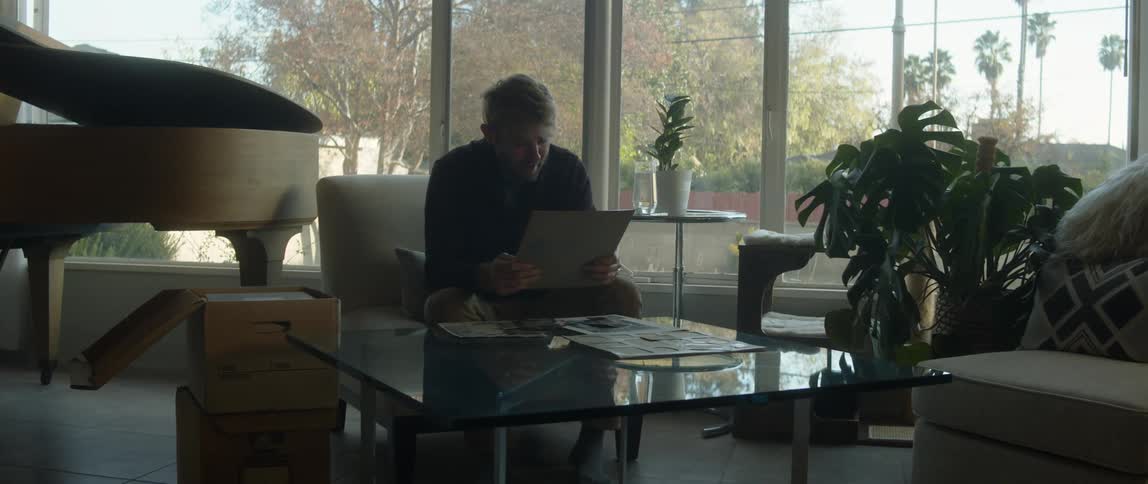 A man looking at photographs in a cardboard box while sitting on a living room chair