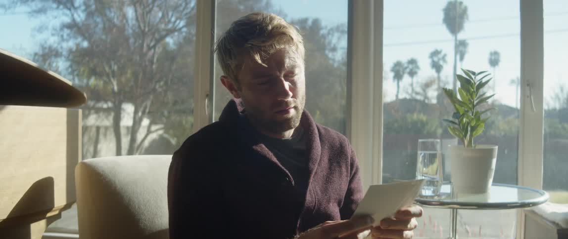 A man is sitting near windows in his living room and reading papers