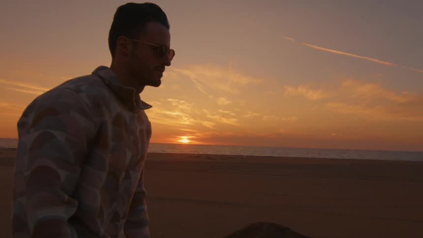 A man is riding a skateboard on a road along the beach with the sun setting in the distance