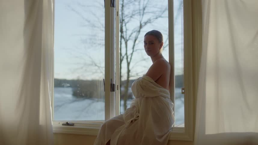 Young woman enveloped in gauze sitting on the windowsill looking at the camera at winter