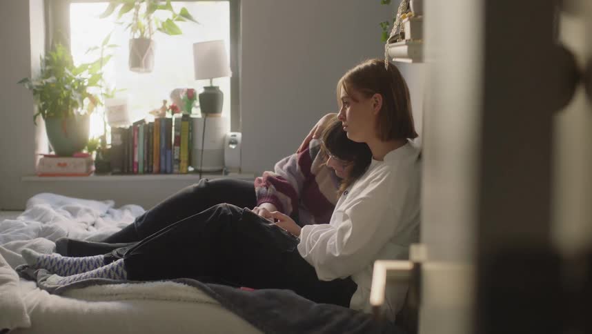 Two teenage girls are sitting on a bed and one girl has her arm around the other comforting her