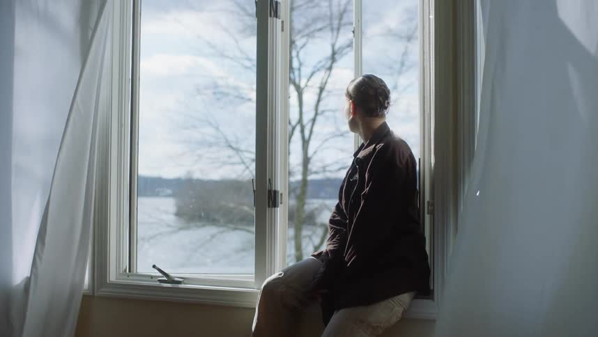 A woman is sitting inside on the windowsill of an open window as she looks outside