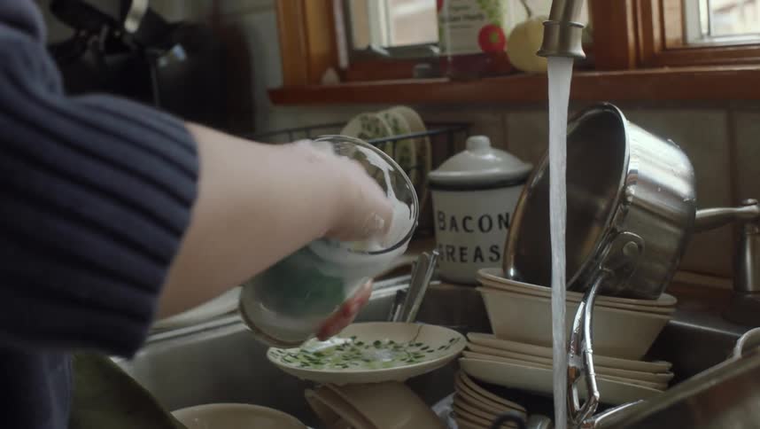 A woman picks up a glass and washes it in a sink full of dirty dishes