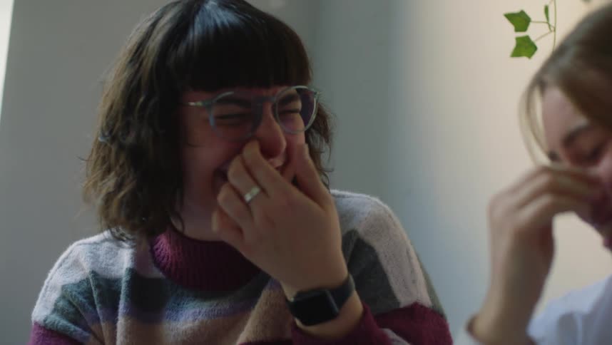 Two women are sitting in a room covering their mouths as they laugh