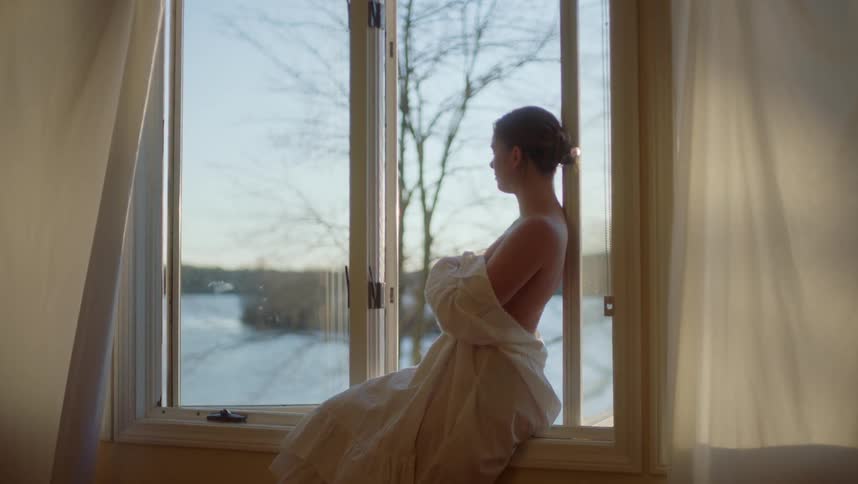A topless woman wrapped in a sheet sitting on a window sill