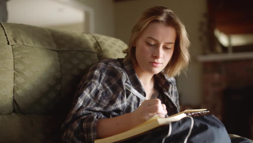 A woman is sitting on her living room couch writing in a book