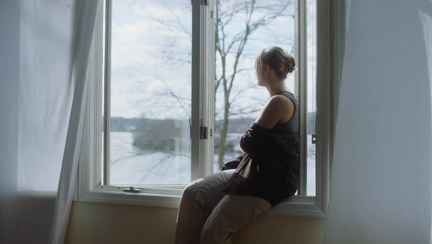 A woman is sitting inside on an open window ledge as she looks outside