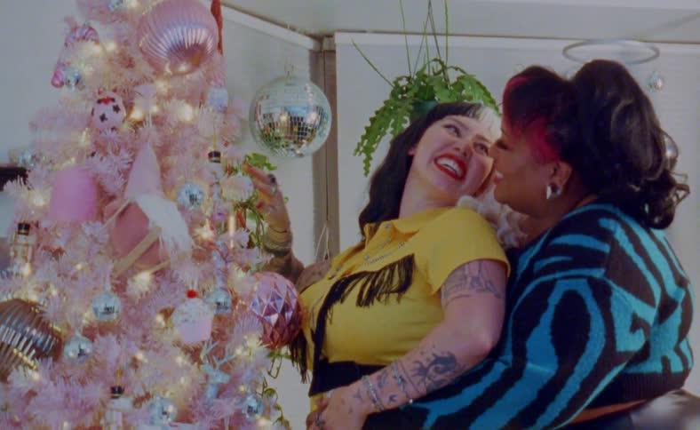 A woman hugs another woman from behind as they laugh while decorating a christmas tree at home