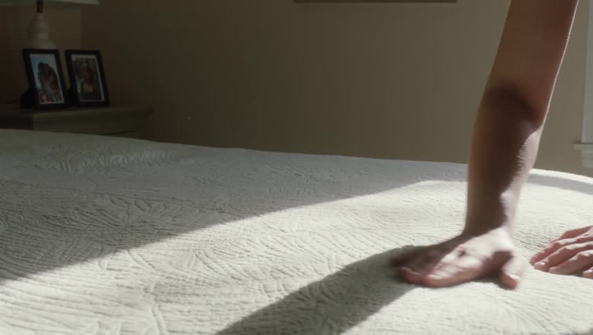 A woman is making her bed and brushing off the sheet and she stands over it looking with her hands on her hips