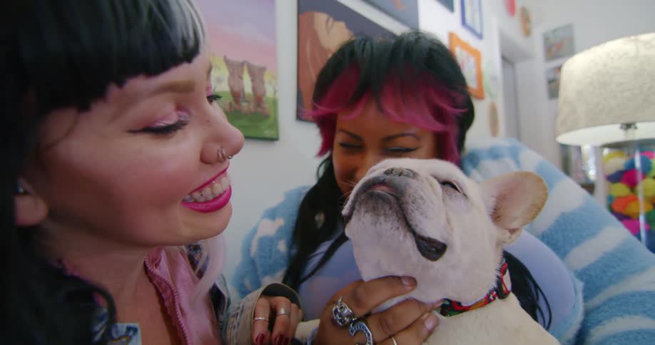 Two female friends pet and nuzzle dog in room decorated with wall art