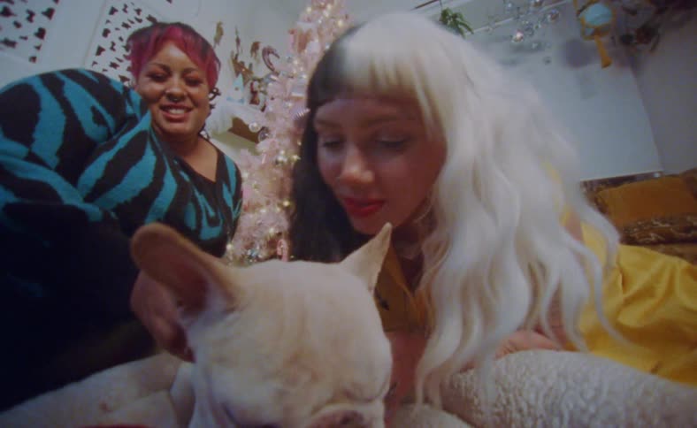 Two women smile while they pet and kiss sleepy dog near christmas tree