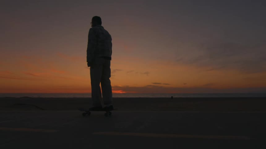 Silhouette of a man riding a skateboard on a road along the beach with the sun setting on the horizon