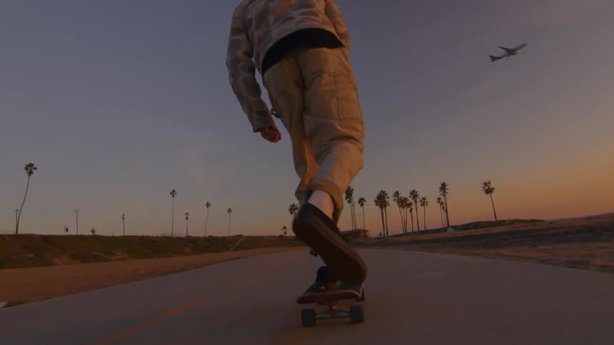 A man is riding a skateboard on a road along the beach as an airpline flies across the sky and there are palm trees in the distance