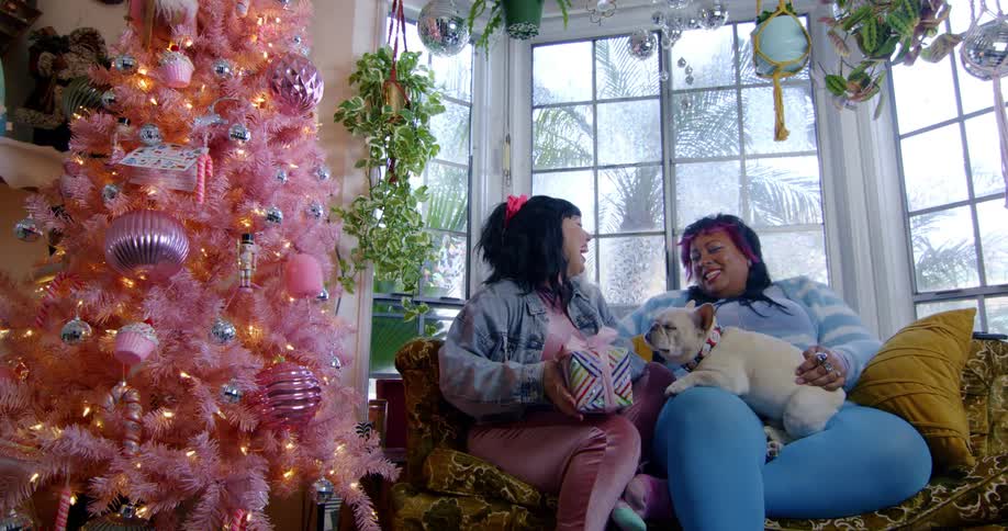 Two women sit on a couch with a dog near a pink christmas tree and one of the women holds up gifts