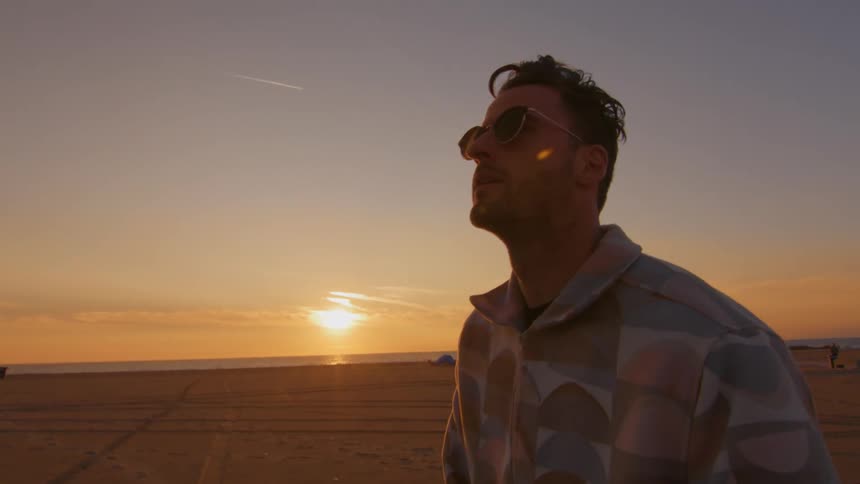 A man is riding a skateboard on a road along the beach as the sun is setting in the distance