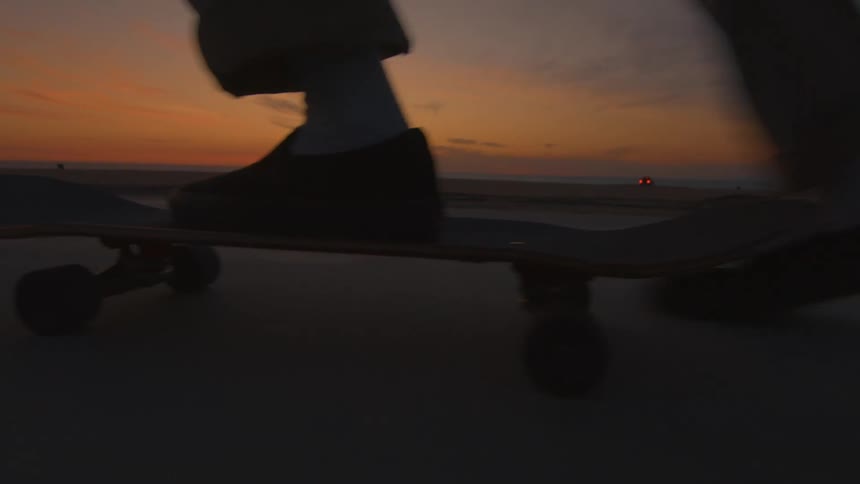 View of a mans feet as he is riding a skateboard on a road along the beach with the sun setting in the distance