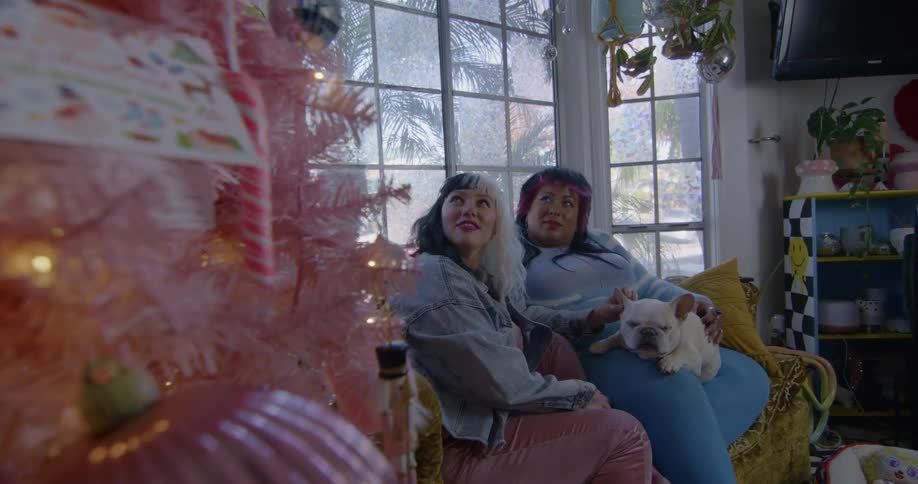 Two women sit on a couch in a house and smile as they look at a pink christmas tree