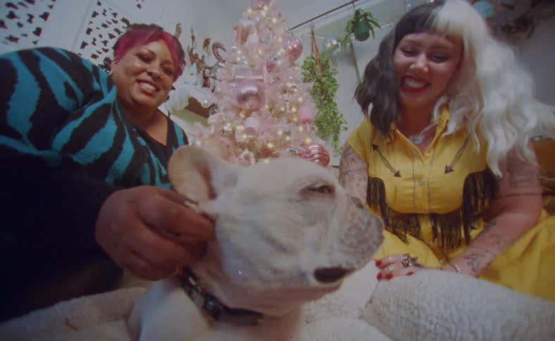 Two women sit near christmas tree petting dog before sharing a kiss