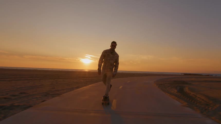 A man is riding a skateboard on a road along the beach with the sun setting in the distance