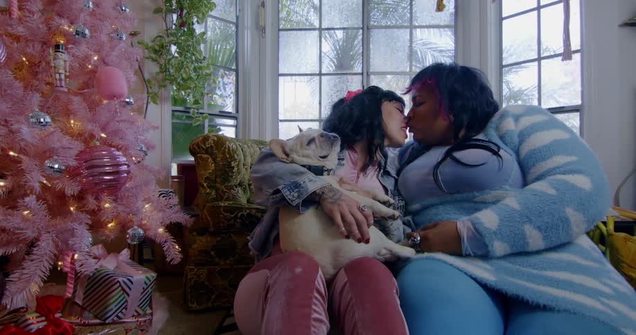 Two women with a dog kiss as they sit in front of a holiday tree