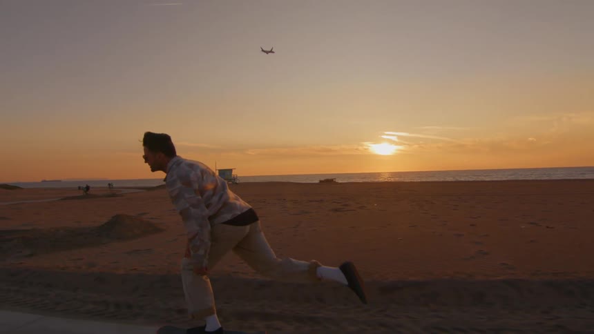 A man is riding a skateboard on a road along the beach as an airplane flies near the sun setting on the horizon