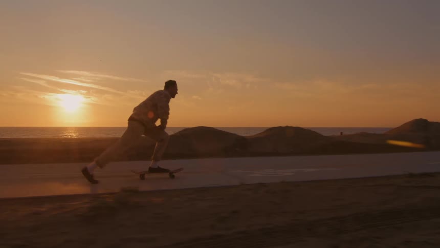 A man is riding a skateboard on a road along the beach as the sun is setting in the distance