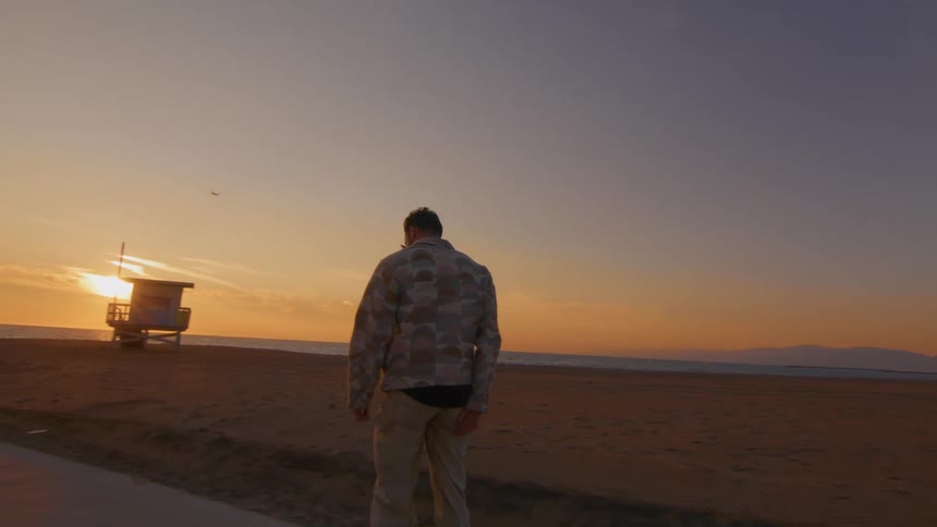 A man is riding a skateboard on a road along the beach as the sun is setting on the horizon