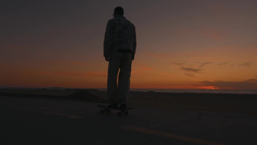 Silhouette of a man riding a skateboard on a road along the beach as the sun is setting on the horizon