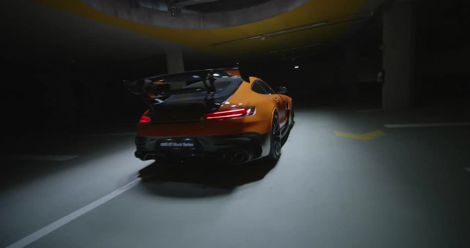 An FPV drone shot zooming around an orange luxury sports car parked under a spotlight inside a dark parking garage
