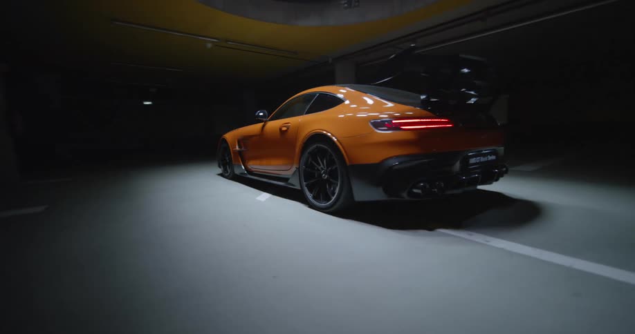 An FPV drone shot of an orange luxury sports car parked under a spotlight inside a dark parking garage
