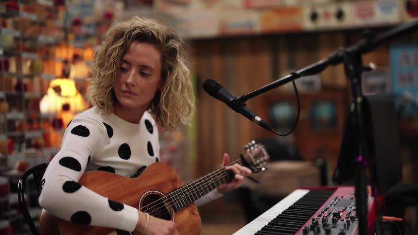 A woman is sitting behind a keyboard and microphone playing an acoustic guitar