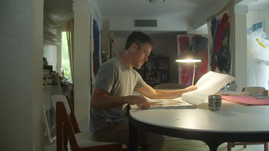 A man at home picks up a book and takes it to a table and flips through it