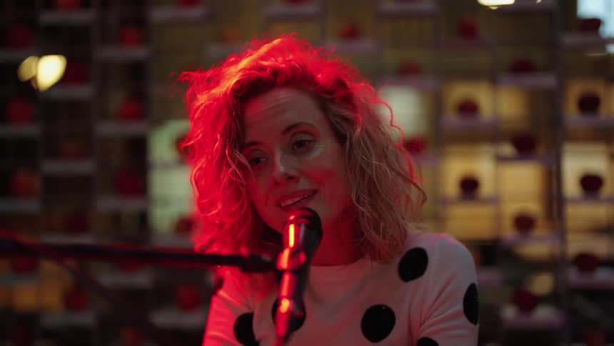Light shines on a woman as she is singing into a microphone and playing a keyboard