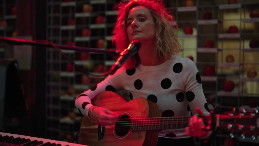 Blonde woman with wavy hair and white spot shirt plays guitar on bar stage